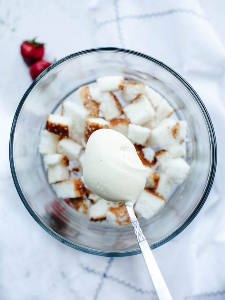 whipped cream being spread on top of angel food cake pieces in a trifle bowl.