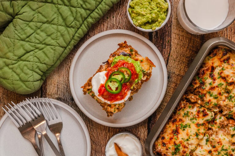 A plate with a slice of cheesy lasagna topped with sour cream, jalapeños, and salsa, alongside avocado dip and a glass of milk.