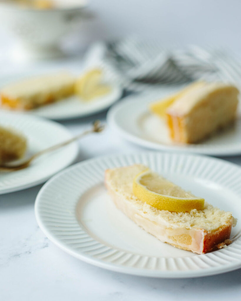 a slice of lemon quick bread on a white plate with a lemon wedge on top.