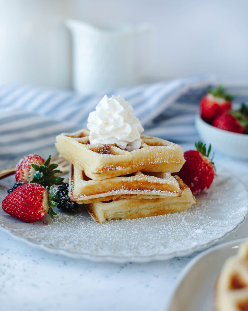 fluffy waffle recipe stacked on a plate with berries and whipped cream.