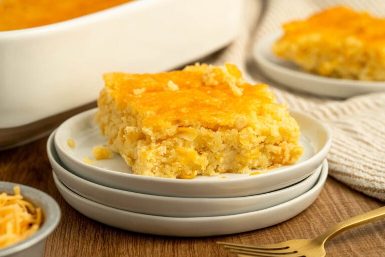 A square piece of cornbread with a golden-brown top sits on a stack of white plates. The cornbread has a fluffy texture with visible corn kernels. In the background, there is a baking dish with more cornbread and another piece on a separate plate. A gold fork and a small bowl of grated cheese are also visible.