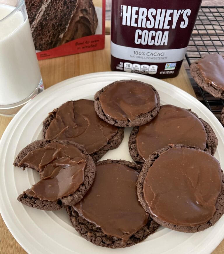 Chocolate cookies topped with spreads on white plate with cocoa powder and milk nearby.