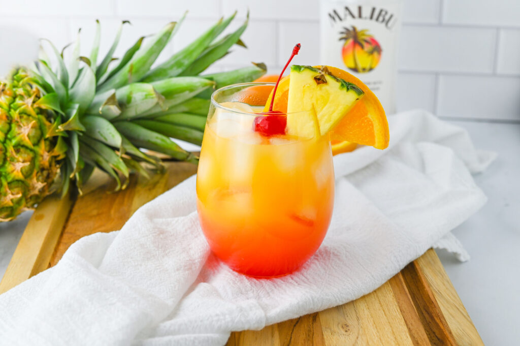 a malibu sunset cocktail drink in a glass with pineapple and malibu rum behind it.