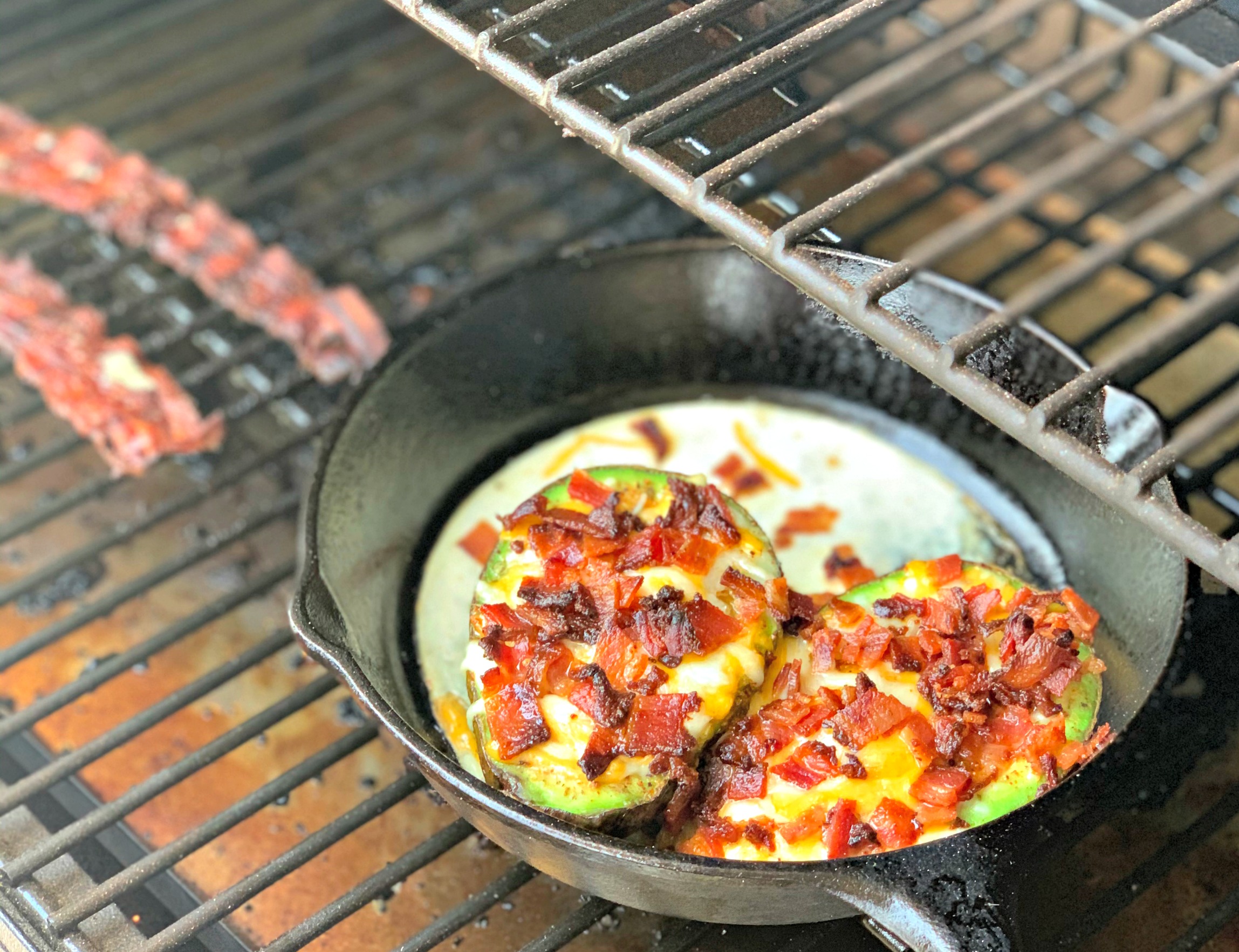 bacon, egg, and cheese stuffed avocados cooking in a cast iron skillet on the grill.