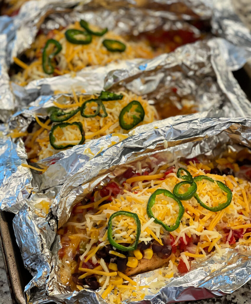 foil packets lined up with chicken, cheese, beans, jalapenos, and tomatoes ready to be grilled