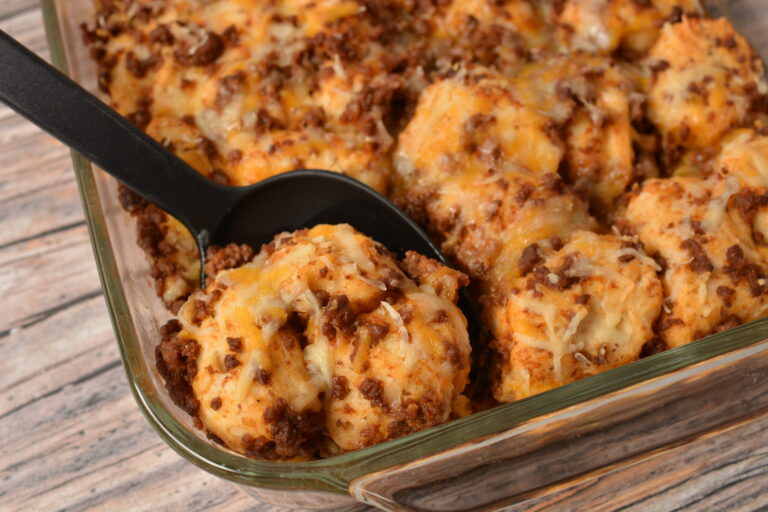 A close-up of cheesy, baked casserole with ground beef and biscuit pieces, served with a black spoon in a glass dish on a wooden table.