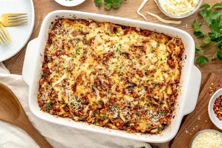 A baked pasta dish topped with melted cheese and garnished with herbs, served in a white casserole dish on a wooden table.