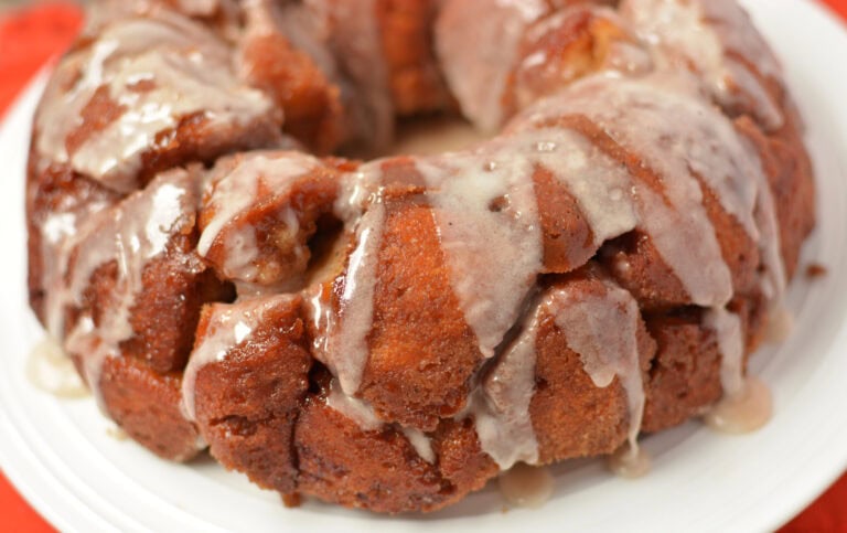 A close-up view of a round, glazed monkey bread on a white plate. The bread is golden brown with a sticky, sugary glaze dripping over the pieces, creating a shiny and tempting appearance.