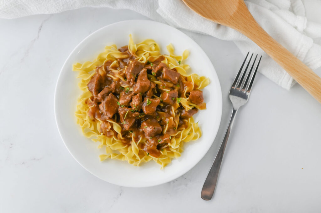 tender beef tips served over egg noodles on a white plate.