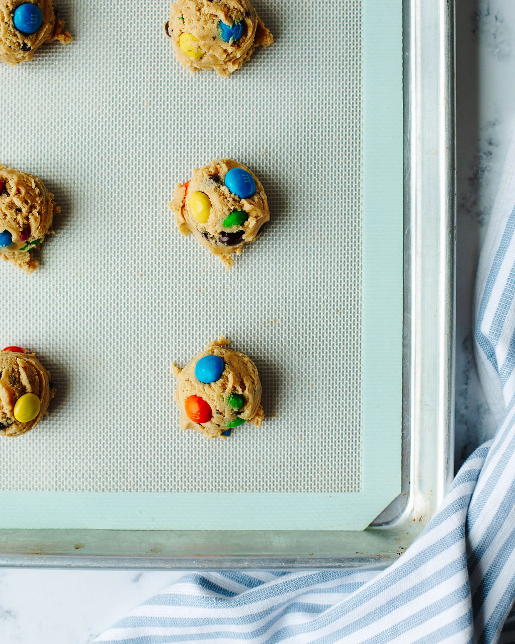 Cookie dough balls topped with peanut butter M&Ms ready to bake on a silicone lined baking sheet.