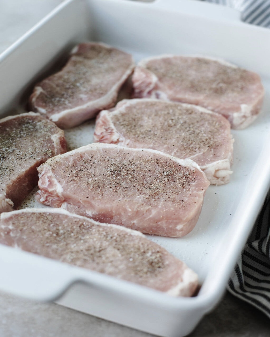 salt and pepper seasoned pork chops in a white baking dish.