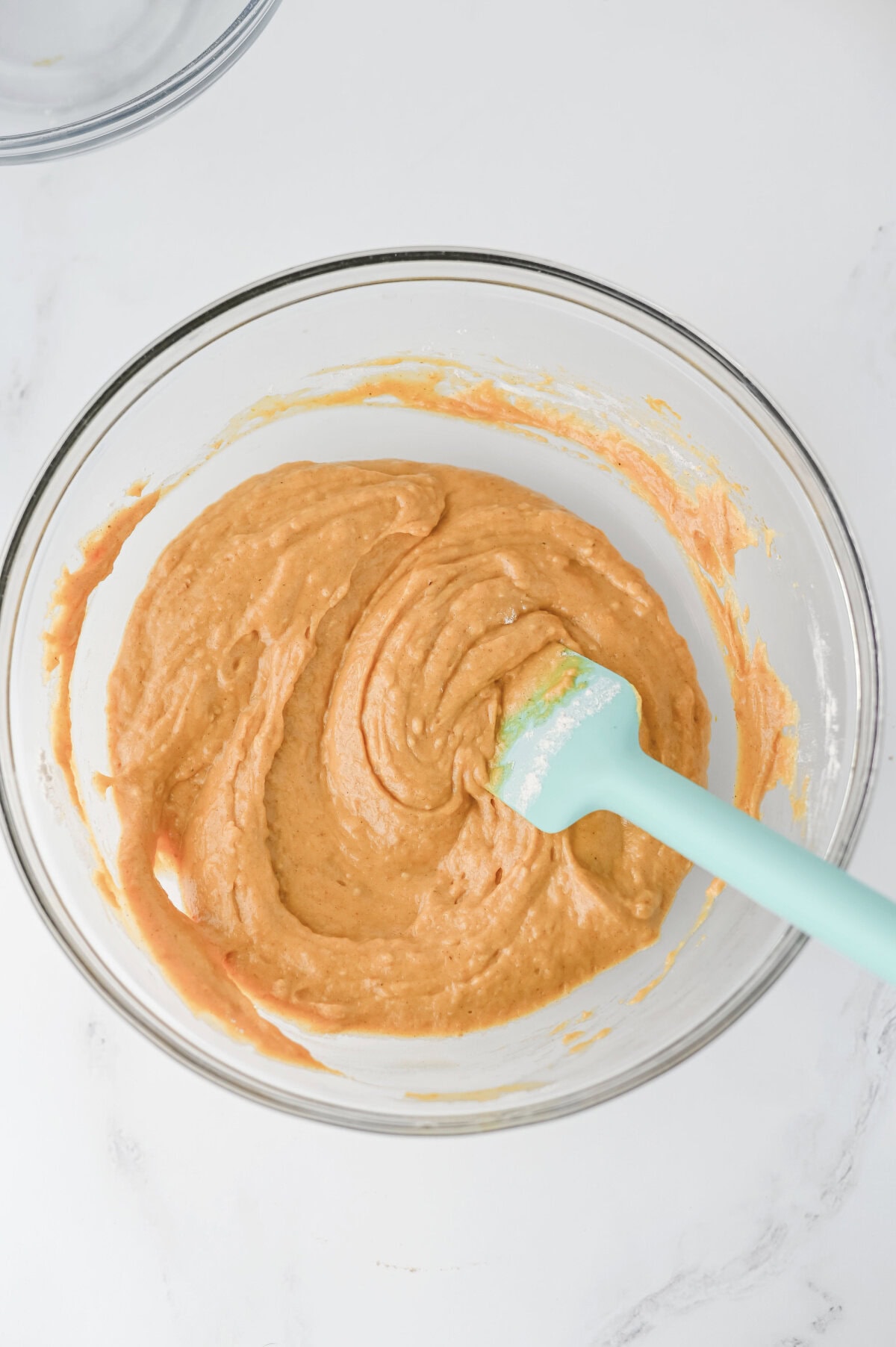 a glass mixing bowl with pumpkin batter combined with a rubber spatula.