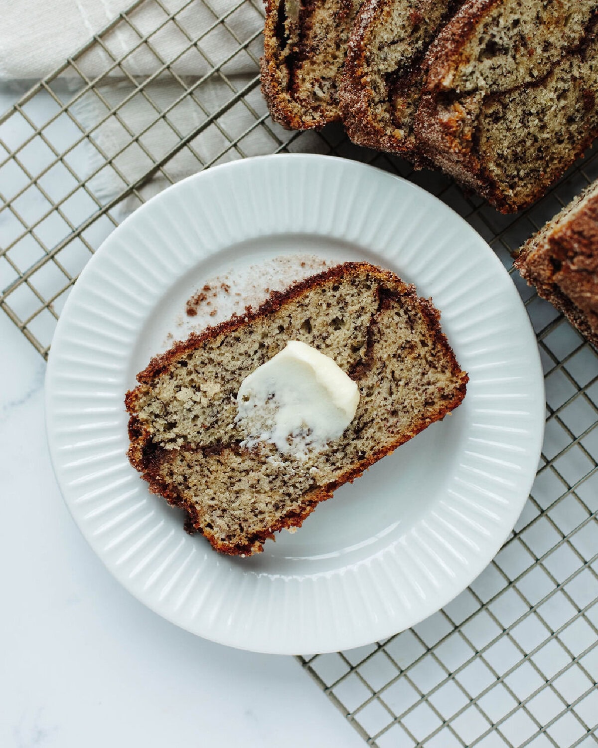 a slice of banana bread with a snickerdoodle swirl throughout on a white plate with a spread of butter.