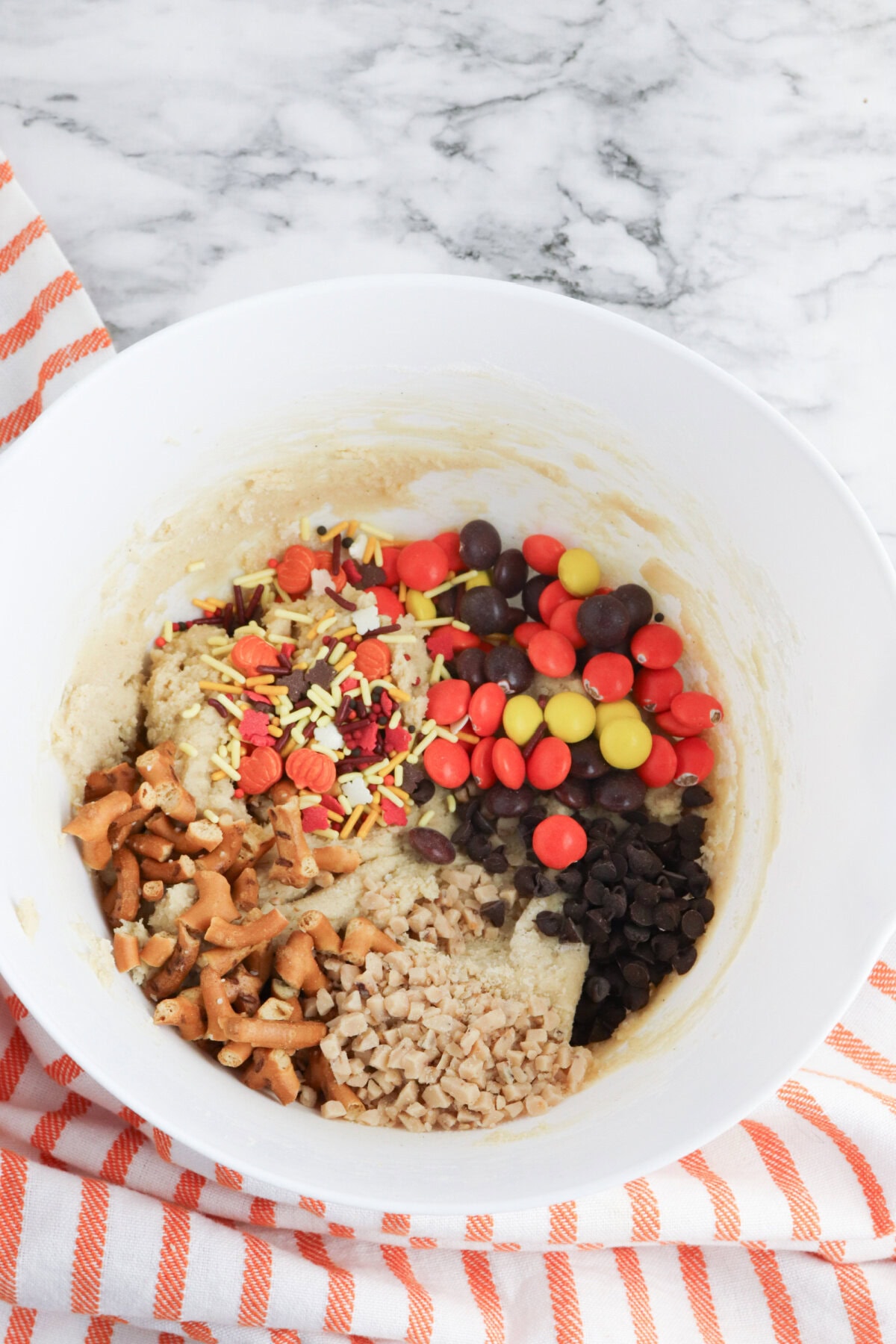 crushed pretzels, reese pieces, sprinkles, and toffee bits added to cookie dough in a mixing bowl.