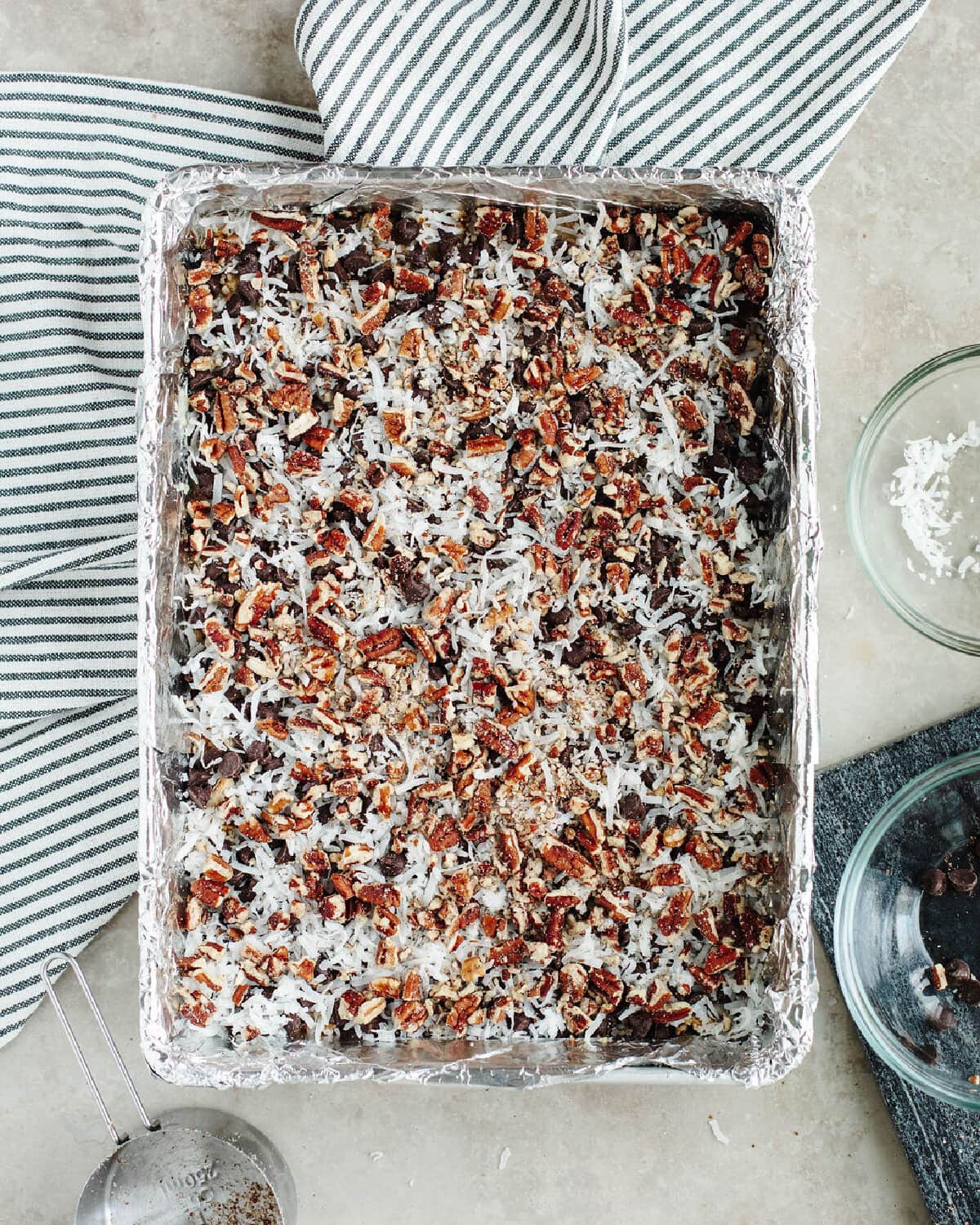 shredded coconut, chopped nuts, and chocolate chips on top of a graham cracker crust in a foil lined baking pan.