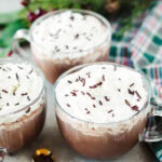 glass mugs of homemade hot cocoa topped with whipped cream and sprinkles.