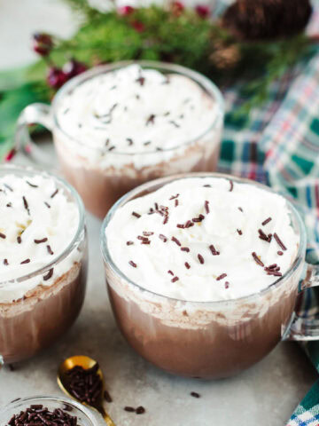 glass mugs of homemade hot cocoa topped with whipped cream and sprinkles.