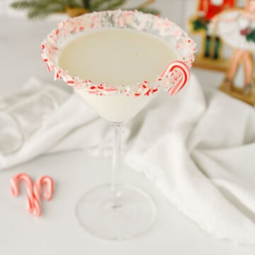 a festive candy cane rimmed martini glass filled with white chocolate peppermint martini and garnished with a mini candy cane.