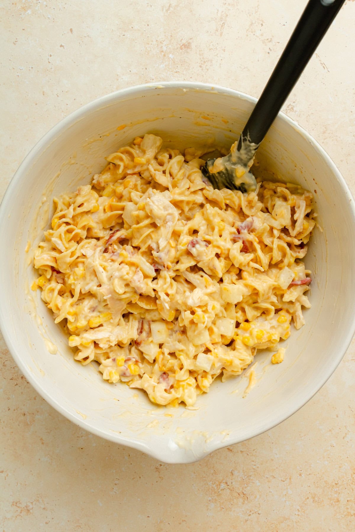 Cooked pasta, chicken, corn, bacon, cheese, and cream of chicken soup combined in a mixing bowl with a spoon.