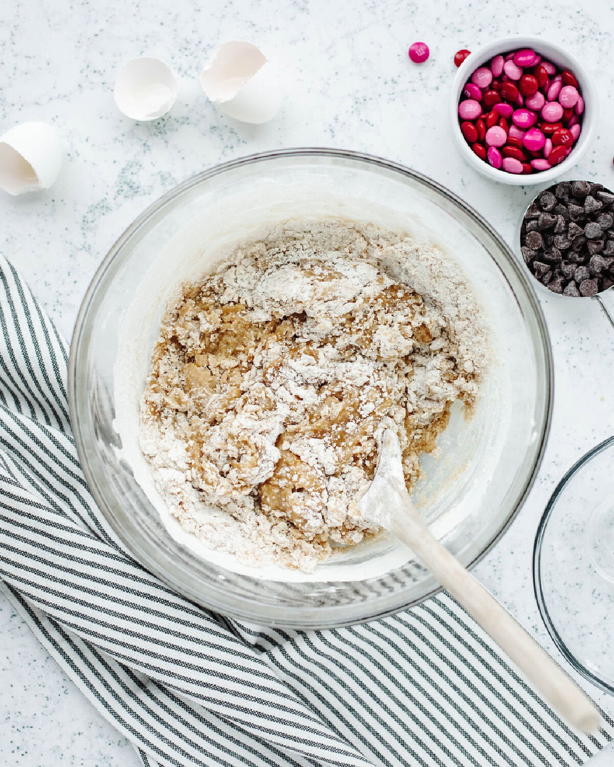 Flour, baking soda, and salt added to cookie dough in a glass mixing bowl using a wooden spoon.