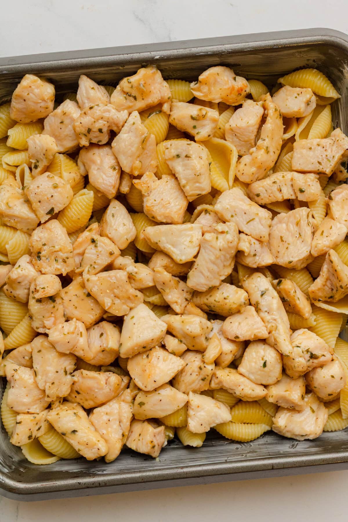 Seasoned diced chicken over cooked pasta in an 8x8 baking pan.