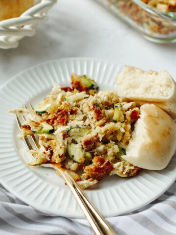 A serving of chicken zucchini casserole with stuffing on a white plate with a dinner roll.