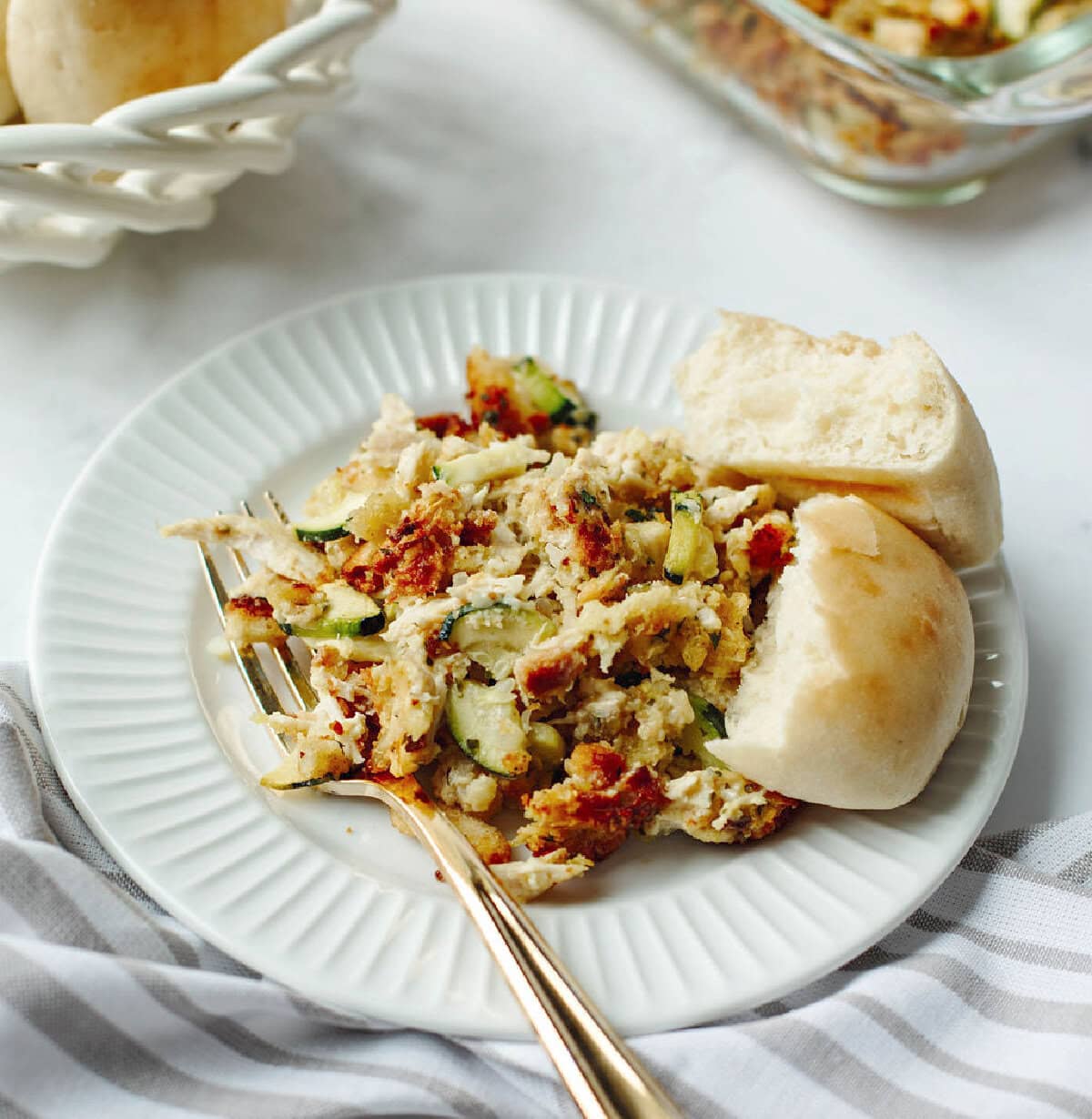 A serving of chicken zucchini casserole with stuffing on a white plate with a dinner roll.