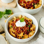Two white bowls with fritos, chili, sour cream, and shredded cheese.