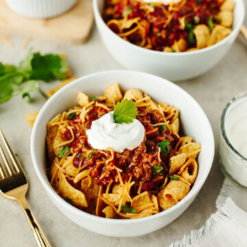 Two white bowls with fritos, chili, sour cream, and shredded cheese.