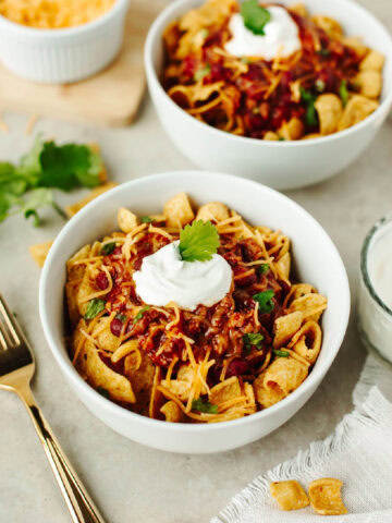 Two white bowls with fritos, chili, sour cream, and shredded cheese.