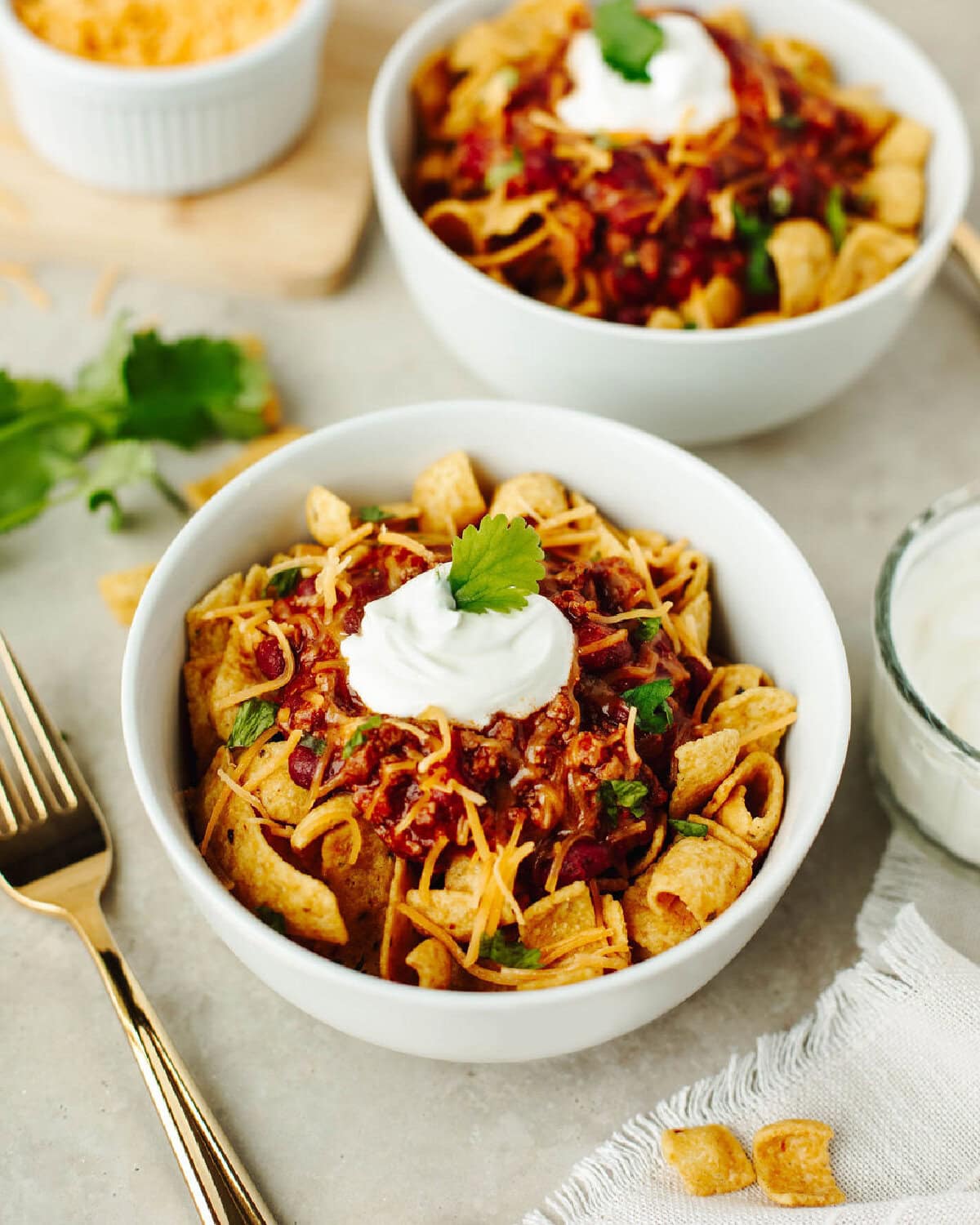 Frito chili pie served in white bowls with sour cream and cheddar cheese.