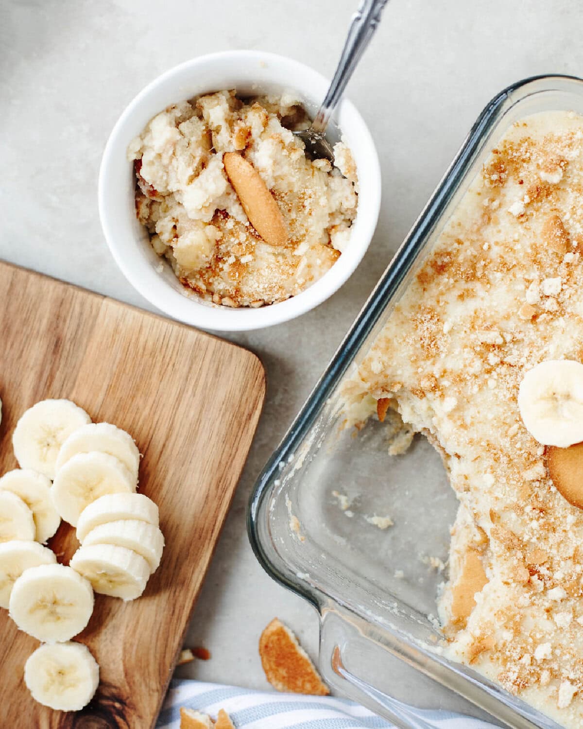 A white dessert bowl of banana pudding with banana slices on a cutting board.