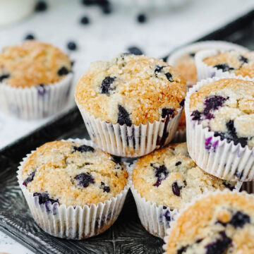 Fresh blueberry muffins with white paper liners stacked on a serving tray.
