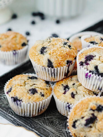 Fresh blueberry muffins with white paper liners stacked on a serving tray.