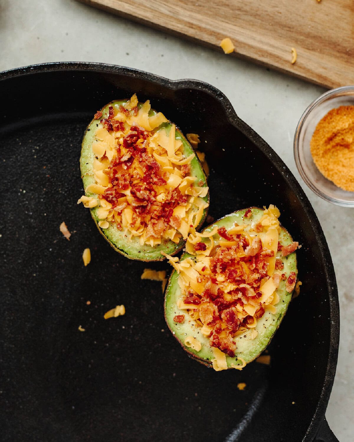 Shredded cheese and crumbled bacon on top of a sliced avocado in a cast iron skillet.