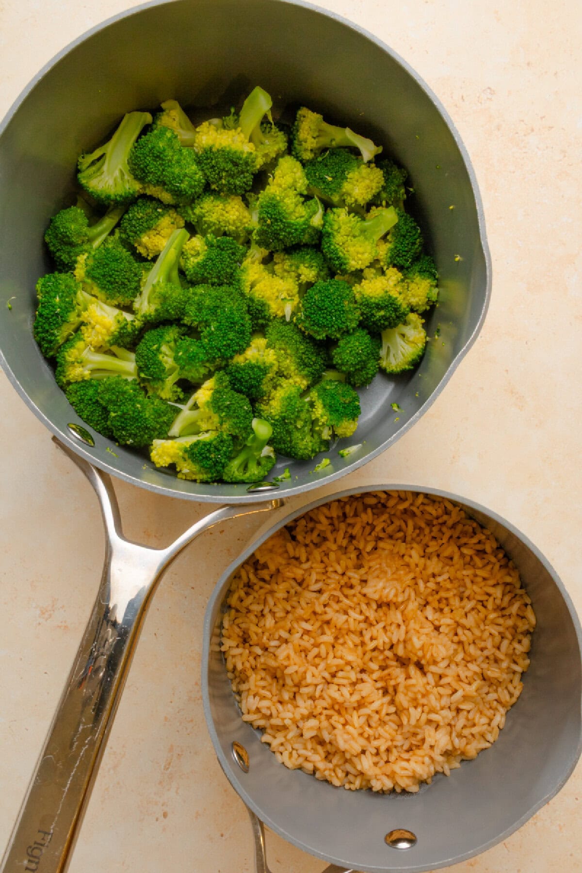 Cooked broccoli in a saucepan and cooked brown rice in a pot.