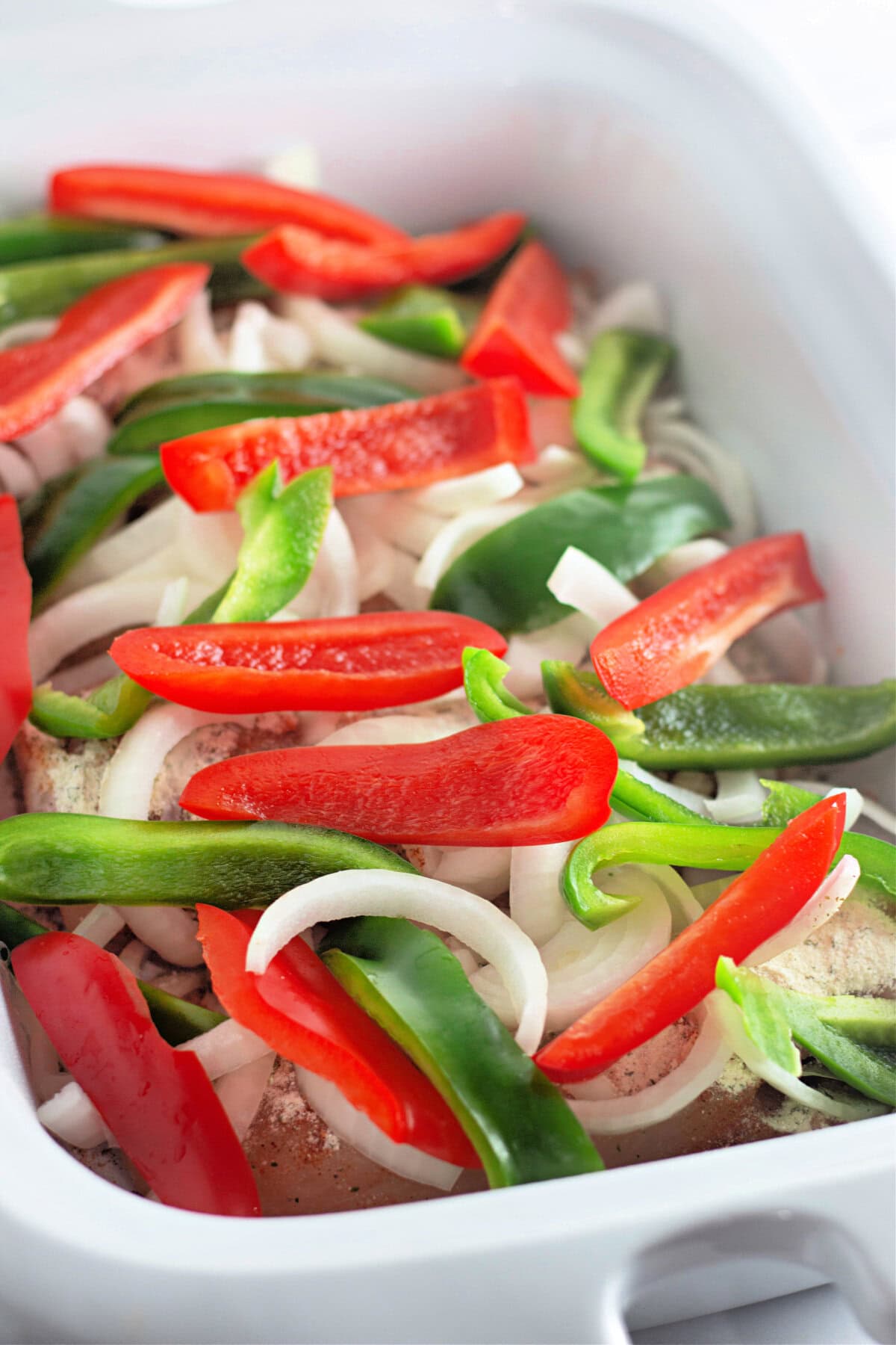 Sliced bell peppers and onion on top of seasoned chicken breasts in a slow cooker.