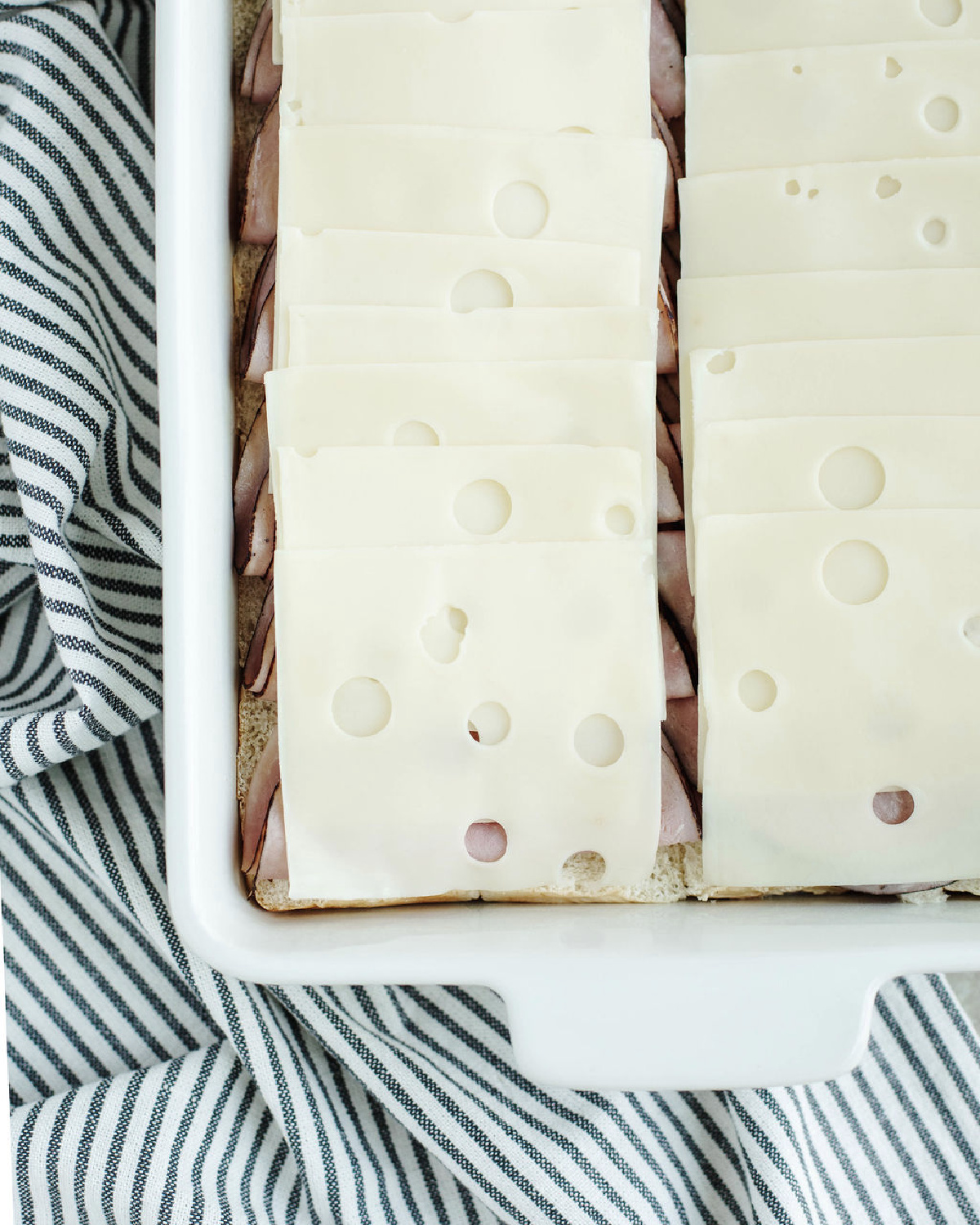 Swiss cheese slices placed on top of ham in a white baking pan.