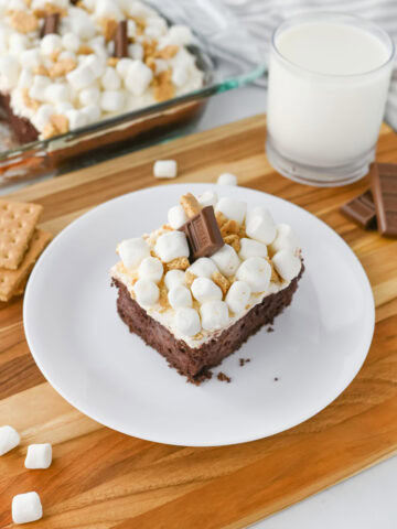 A slice of smores poke cake on a white plate with a glass of milk.