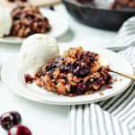Cast iron skillet cherry cobbler on a white plate with a scoop of vanilla ice-cream.