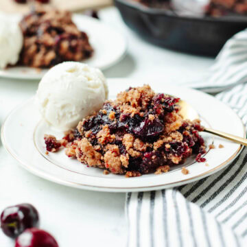 Cast iron skillet cherry cobbler on a white plate with a scoop of vanilla ice-cream.