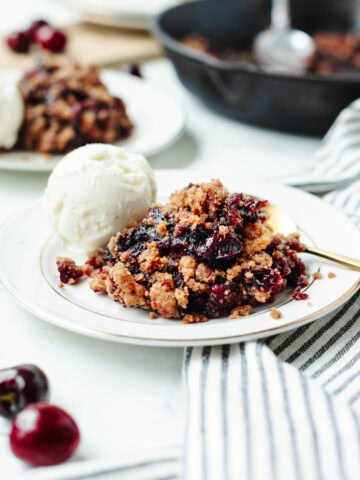 Cast iron skillet cherry cobbler on a white plate with a scoop of vanilla ice-cream.