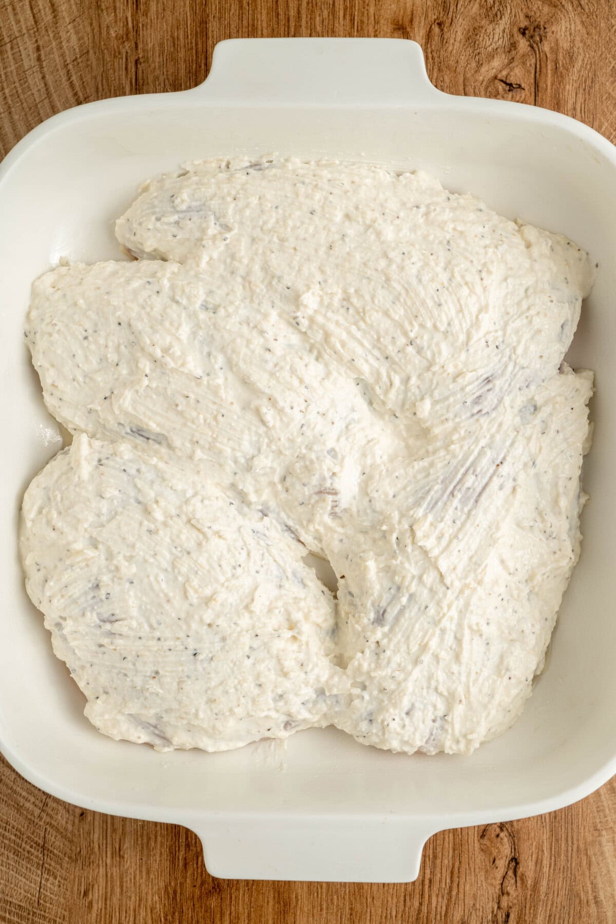 Mayonnaise, Parmesan, salt, and pepper coating chicken breasts in a white casserole dish.