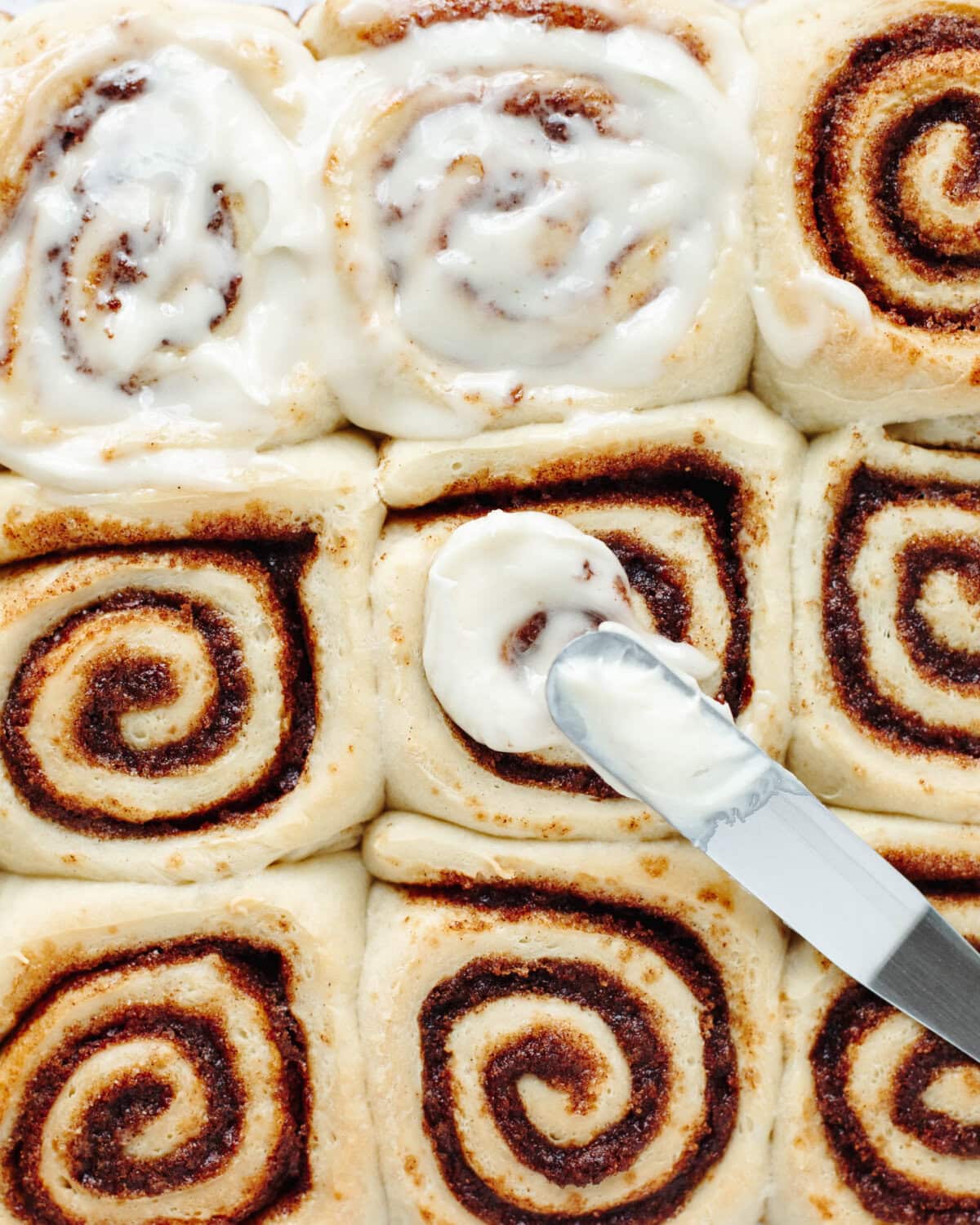 Cream cheese frosting spread using a knife on top of ooey gooey cinnamon rolls.