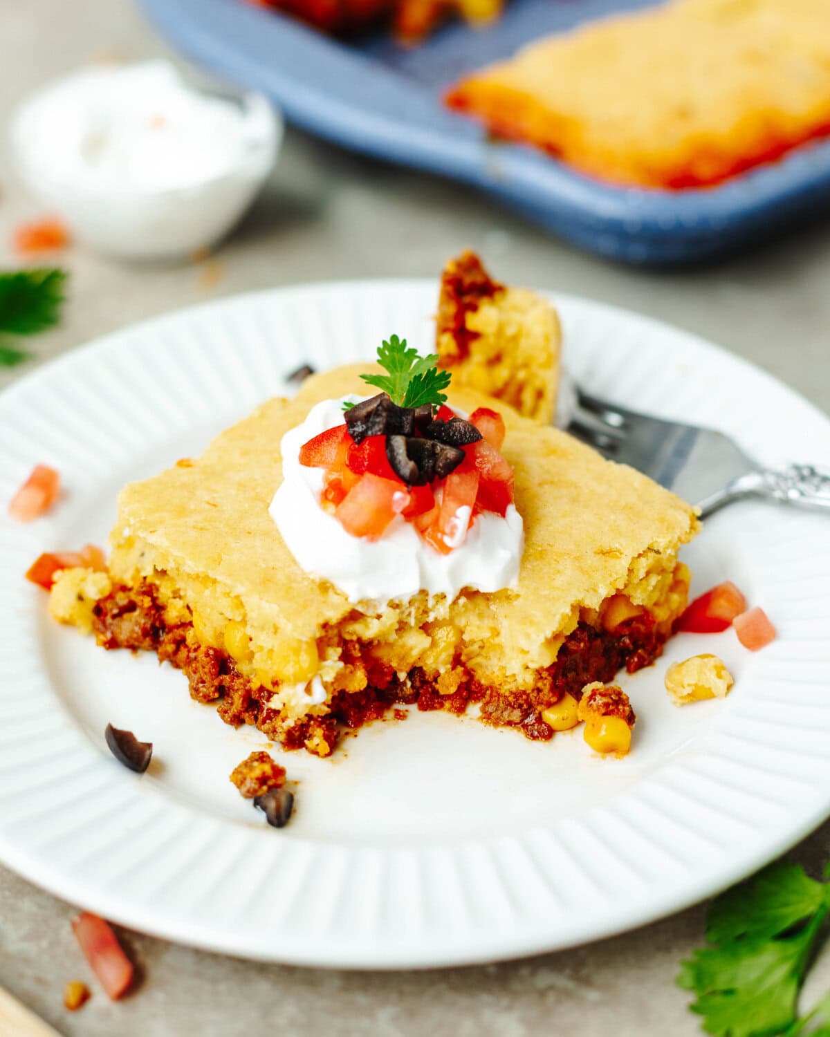 Taco cornbread casserole served on a white plate with sour cream, tomatoes, and olives.