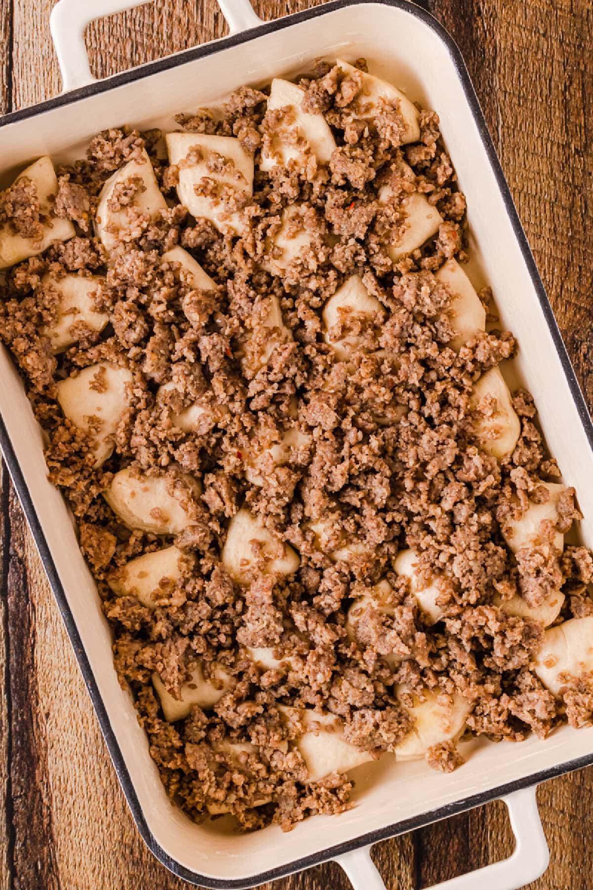 Crumbled sausage on top of quartered biscuits in a baking pan.