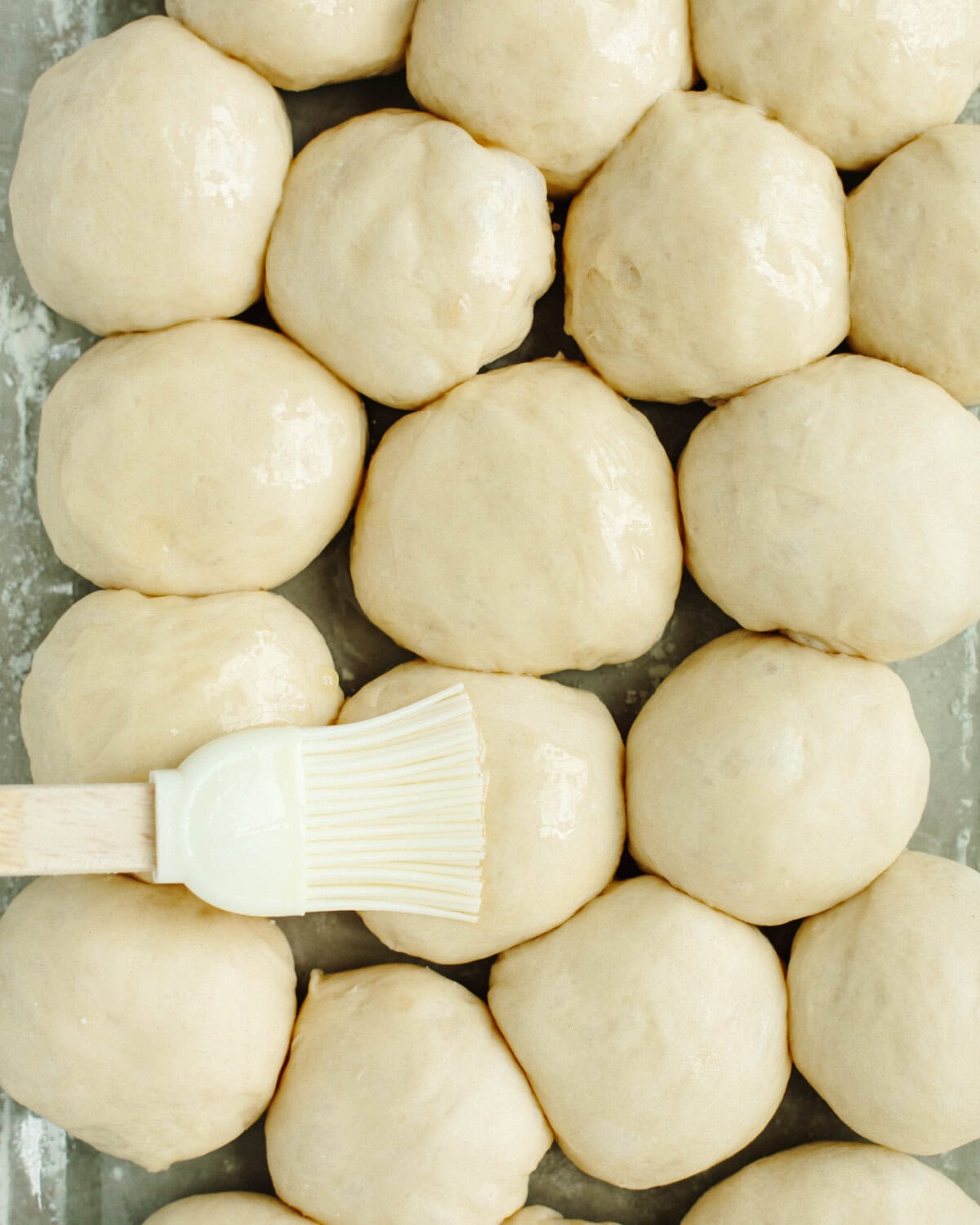 A pastry brush brushing melted butter onto dough balls in a glass baking pan.