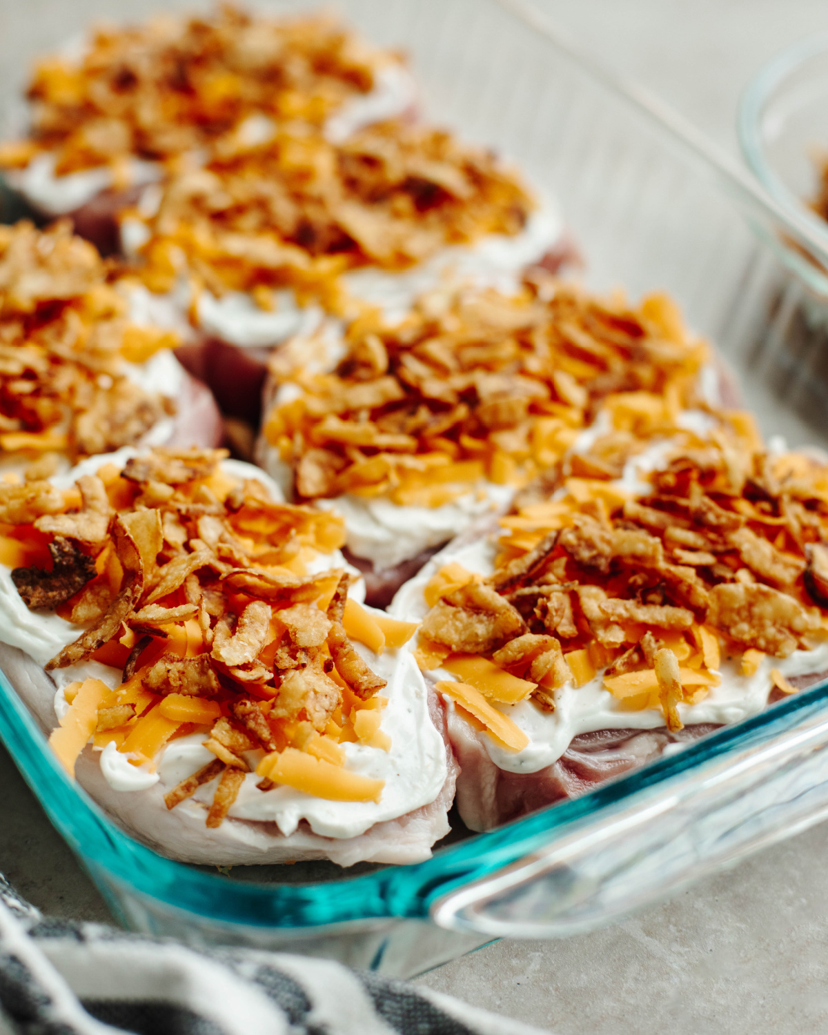 Onion dip, cheddar cheese, and fried onions sprinkled on top of boneless pork chops in a 9x13 baking pan.