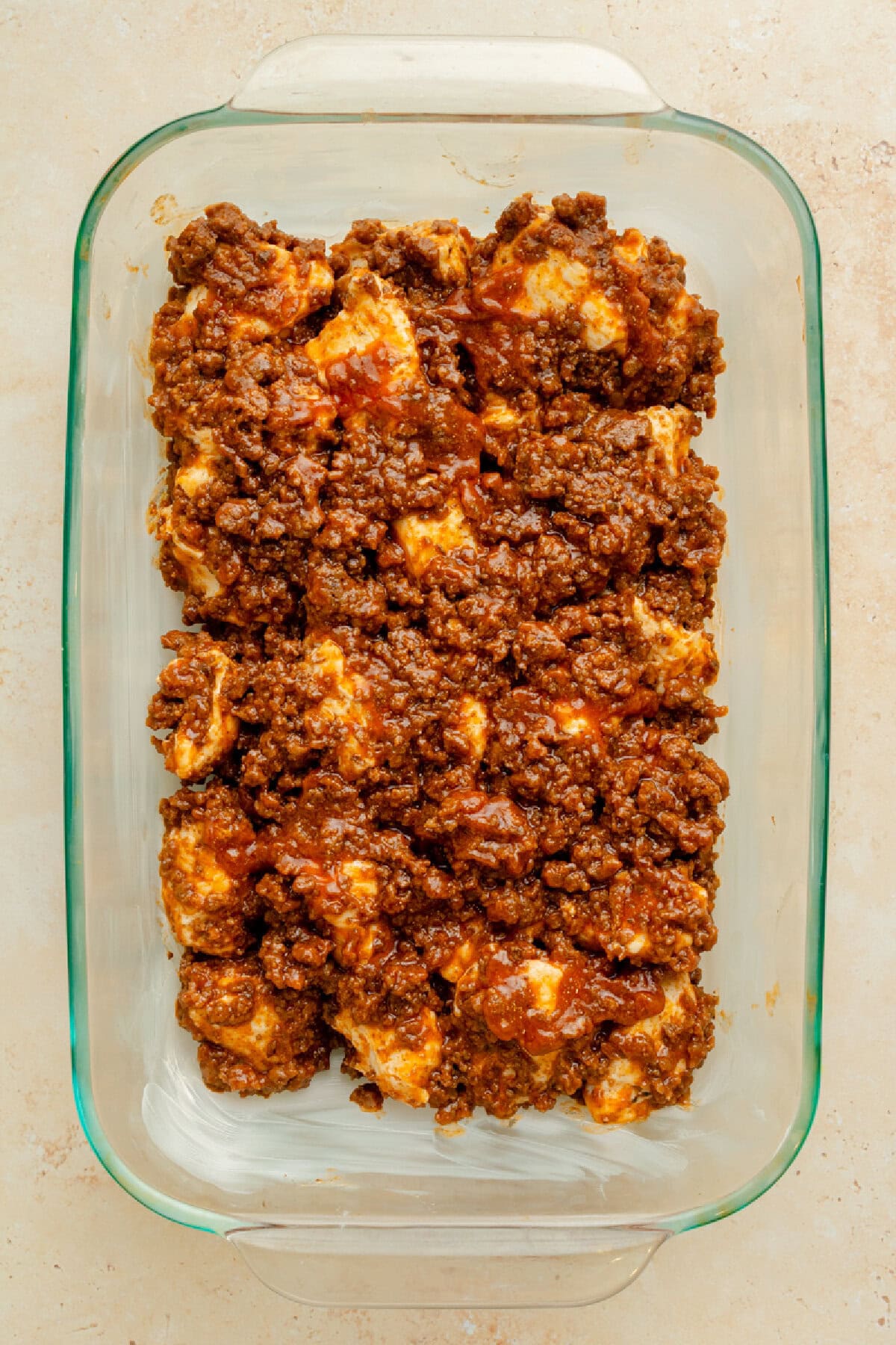 A ground beef and biscuit mixture spread evenly into a prepared 9x13 glass baking pan.