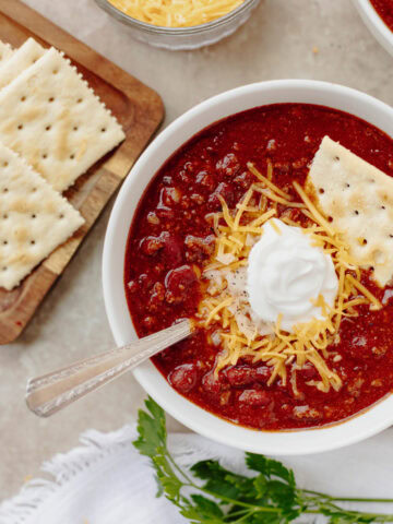 A white bowl with game day chili served with sour cream, crackers, and shredded cheese.
